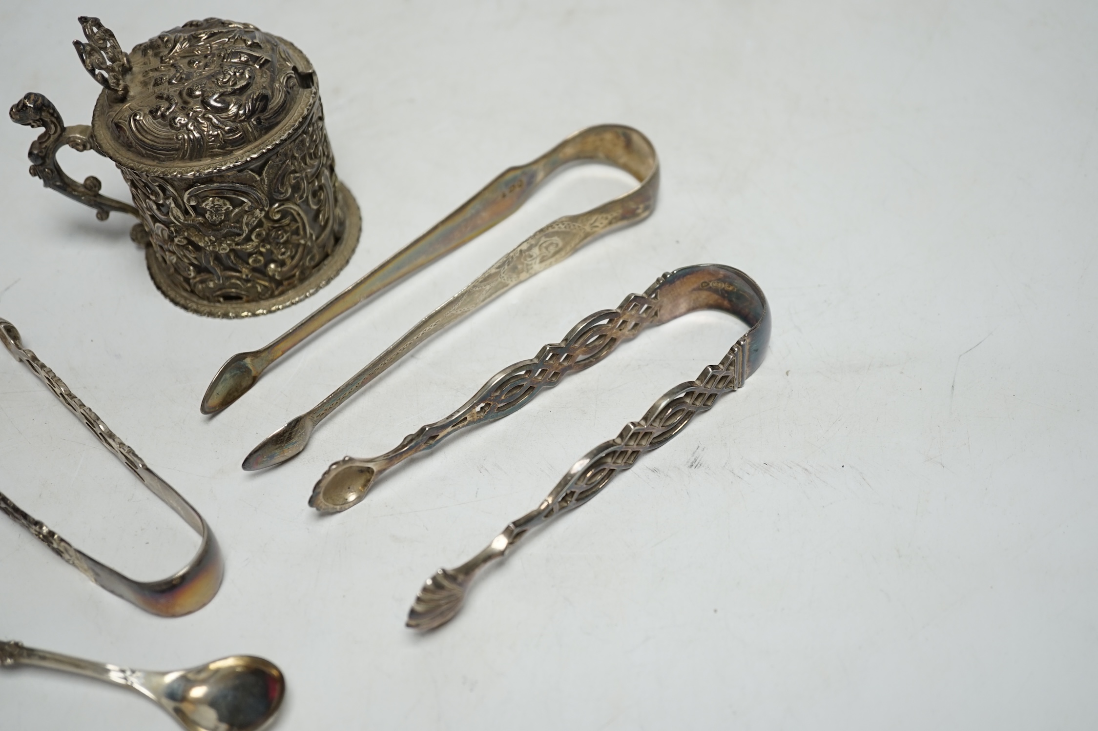 A Victorian silver embossed mustard pot, with spoon and three pairs of George III and later silver sugar tongs.